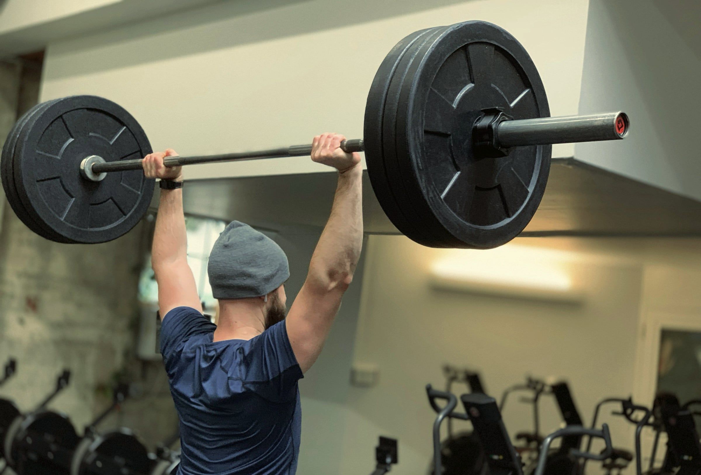 A member in a beanie presses a loaded barbell overhead.