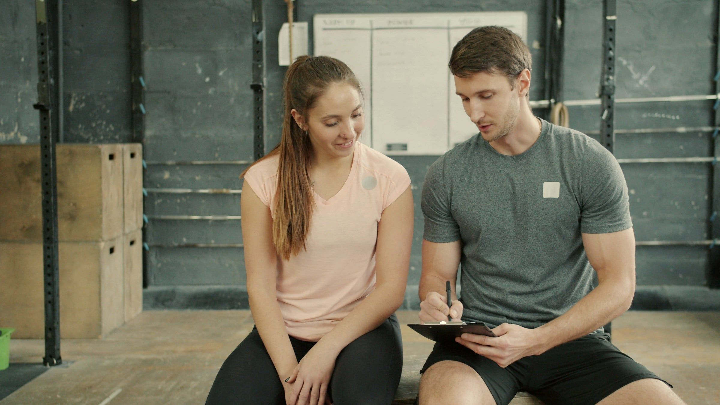 A coach writes notes on a clipboard while his client goes over her session beside him.